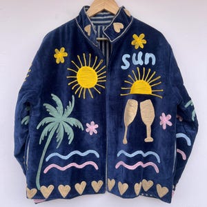 Könnte beinhalten: Marineblaue Samtjacke mit bunten tropischen Stickereien. Sie zeigt eine gelbe Sonne, eine Palme und das Wort "sun" in Blau. Goldene Herzen und Sektgläser sind ebenfalls gestickt. Die Jacke hat ein gestreiftes Futter.