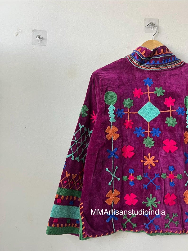 May include: A deep purple velvet jacket with colorful embroidered floral and geometric designs. The jacket features a high collar and long sleeves with detailed embroidery. The text "MMArtisanstudioindia" is visible on the lower right side.