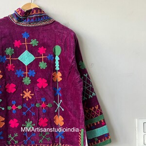 May include: A deep purple velvet jacket with colorful embroidered floral and geometric designs. The jacket features a high collar and long sleeves with decorative trim. The text "MMArtisanstudioindia" is visible on the front of the jacket.