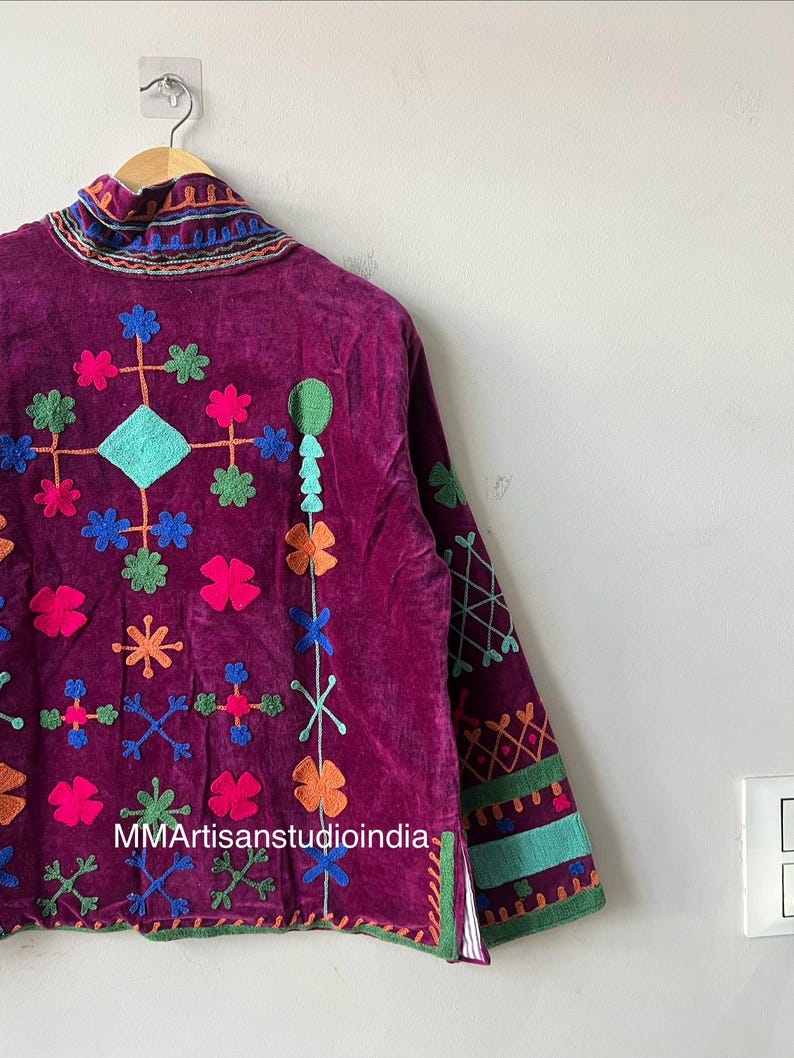 May include: A deep purple velvet jacket with colorful embroidered floral and geometric designs. The jacket features a high collar and long sleeves with decorative trim. The text "MMArtisanstudioindia" is visible on the front of the jacket.