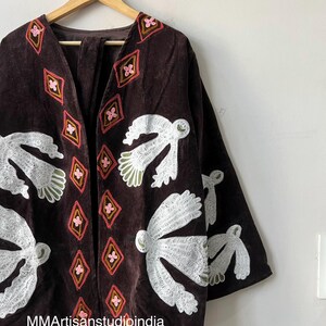 May include: A brown velvet jacket with white bird and pink diamond embroidery. The jacket is hanging on a wooden hanger against a white wall. The text "MMArtisanstudioindia" is visible at the bottom.