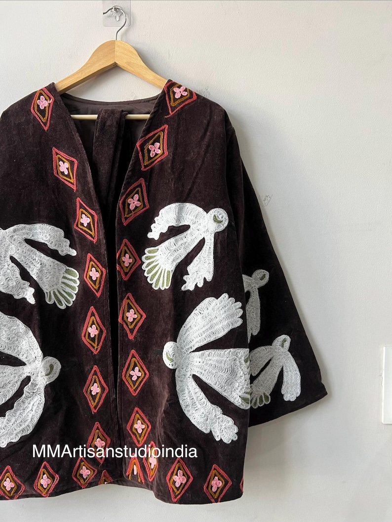 May include: A brown velvet jacket with white bird and pink diamond embroidery. The jacket is hanging on a wooden hanger against a white wall. The text "MMArtisanstudioindia" is visible at the bottom.