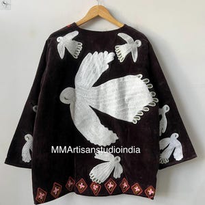 May include: A dark brown, long-sleeved jacket with white embroidered bird designs. The jacket features a round neckline and a decorative hemline with pink and red accents. The text "MMArtisanstudioindia" is visible on the front.