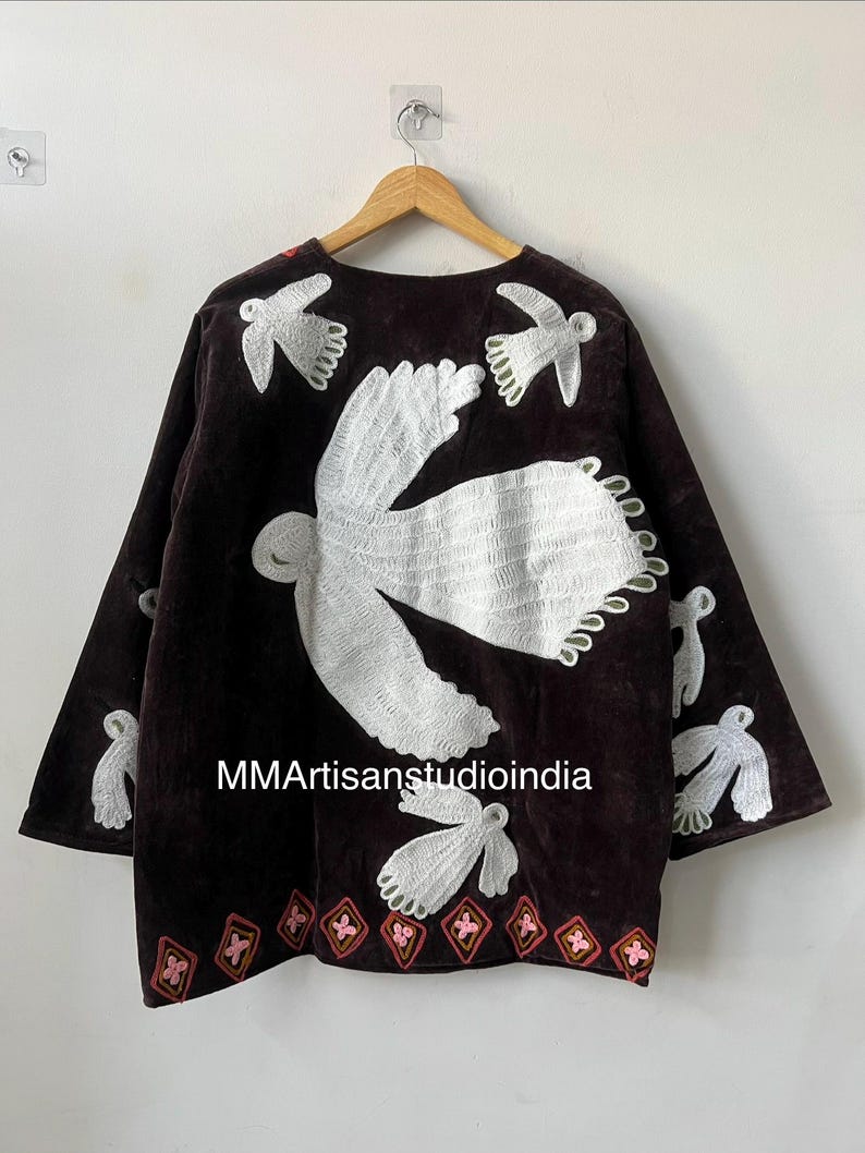 May include: A dark brown, long-sleeved jacket with white embroidered bird designs. The jacket features a round neckline and a decorative hemline with pink and red accents. The text "MMArtisanstudioindia" is visible on the front.