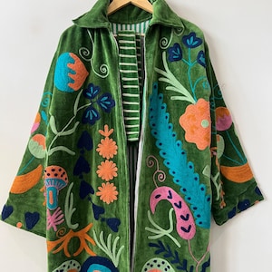May include: A long-sleeved green velvet coat featuring vibrant, multi-colored embroidery of various floral and mushroom motifs. The coat has a collared neckline, a front zipper closure, and a white and green striped lining visible along the inner edges.