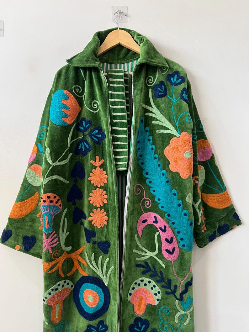 May include: A long-sleeved green velvet coat featuring vibrant, multi-colored embroidery of various floral and mushroom motifs. The coat has a collared neckline, a front zipper closure, and a white and green striped lining visible along the inner edges.