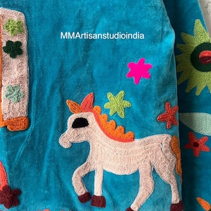 May include: A turquoise textile with embroidered details. A light pink unicorn with an orange mane and tail is featured, surrounded by colorful star and flower accents. The text "MMArtisanstudioindia" is visible.