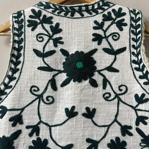 May include: A white vest with dark green floral embroidery. The embroidery features stylized flowers and vines. The vest has a cream-colored, fuzzy lining and is hanging on a wooden hanger. The text "MMArtisanstudioindia" is visible at the bottom.