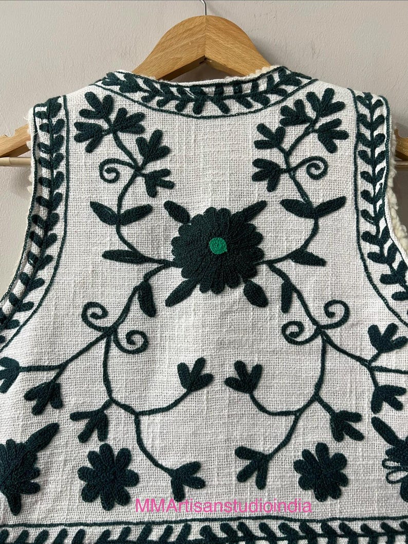 May include: A white vest with dark green floral embroidery. The embroidery features stylized flowers and vines. The vest has a cream-colored, fuzzy lining and is hanging on a wooden hanger. The text "MMArtisanstudioindia" is visible at the bottom.