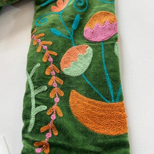 May include: A green velvet fabric featuring dense, colorful embroidery. The design includes stylized floral and botanical motifs in shades of orange, pink, light green, and dark blue, with intricate stitching patterns covering the surface.