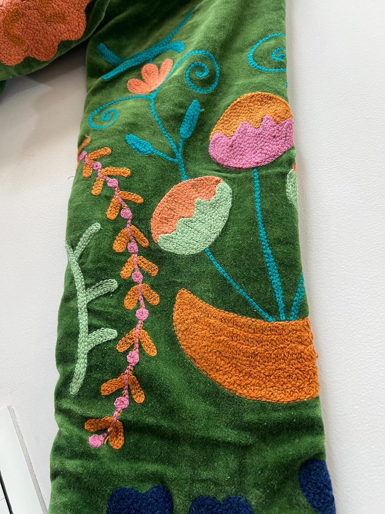 May include: A green velvet fabric featuring dense, colorful embroidery. The design includes stylized floral and botanical motifs in shades of orange, pink, light green, and dark blue, with intricate stitching patterns covering the surface.