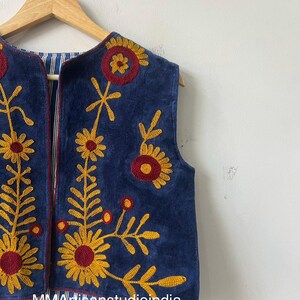 May include: A sleeveless, navy blue vest with intricate floral embroidery in yellow and maroon. The vest is hanging on a wooden hanger against a white wall. The interior lining features a blue and white striped pattern.