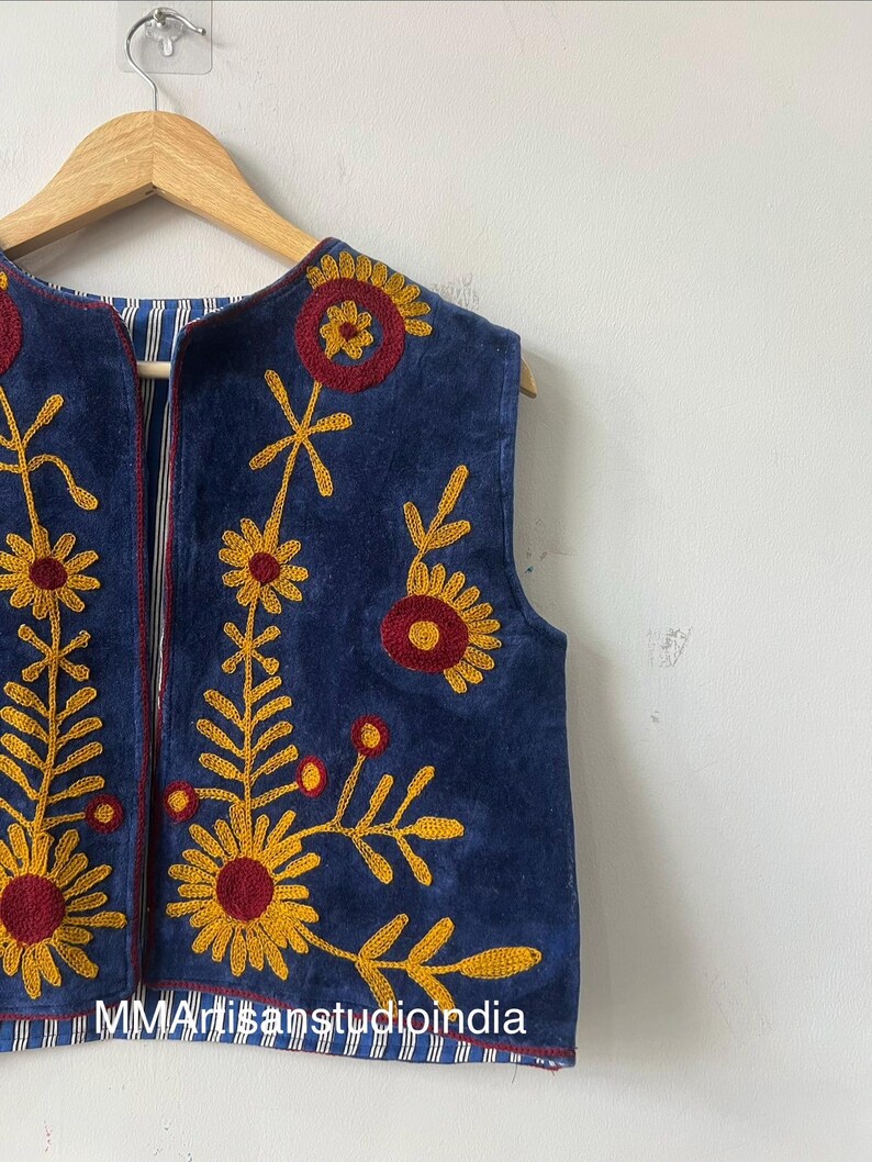 May include: A sleeveless, navy blue vest with intricate floral embroidery in yellow and maroon. The vest is hanging on a wooden hanger against a white wall. The interior lining features a blue and white striped pattern.