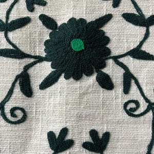 May include: Close-up of a white linen textile with dark green embroidered floral patterns. The design features a central flower with a green center, surrounded by leaves and vines. The text "MMArtisanstudioindia" is at the bottom.