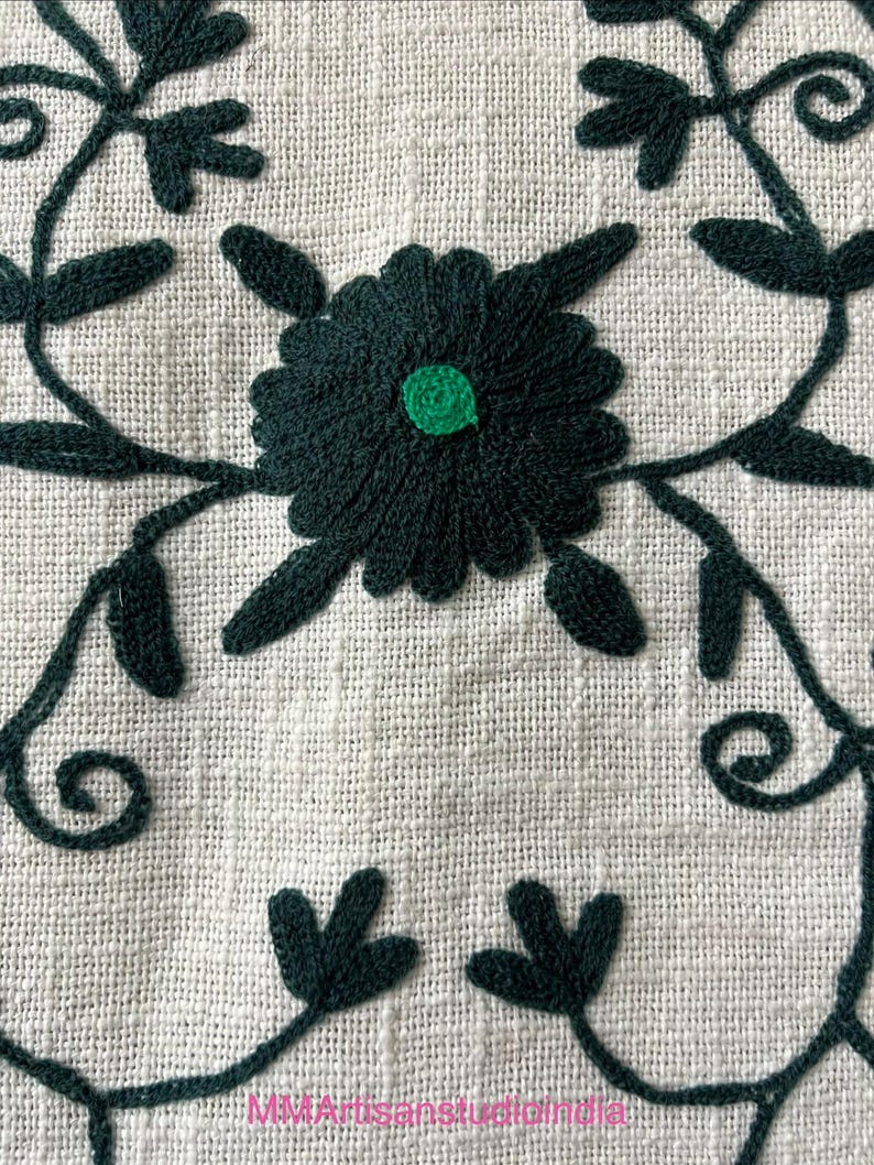 May include: Close-up of a white linen textile with dark green embroidered floral patterns. The design features a central flower with a green center, surrounded by leaves and vines. The text "MMArtisanstudioindia" is at the bottom.
