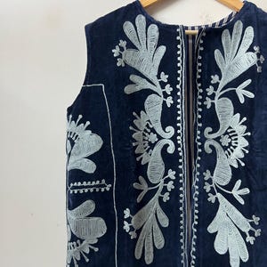Puede incluir: Un chaleco azul marino con intrincados bordados florales plateados. La prenda sin mangas presenta un cuello redondo y una cremallera vertical. El bordado incluye dise&ntilde;os de cachemir y hojas, creando un aspecto detallado y elegante. El chaleco est&aacute; colgado en una percha de madera.