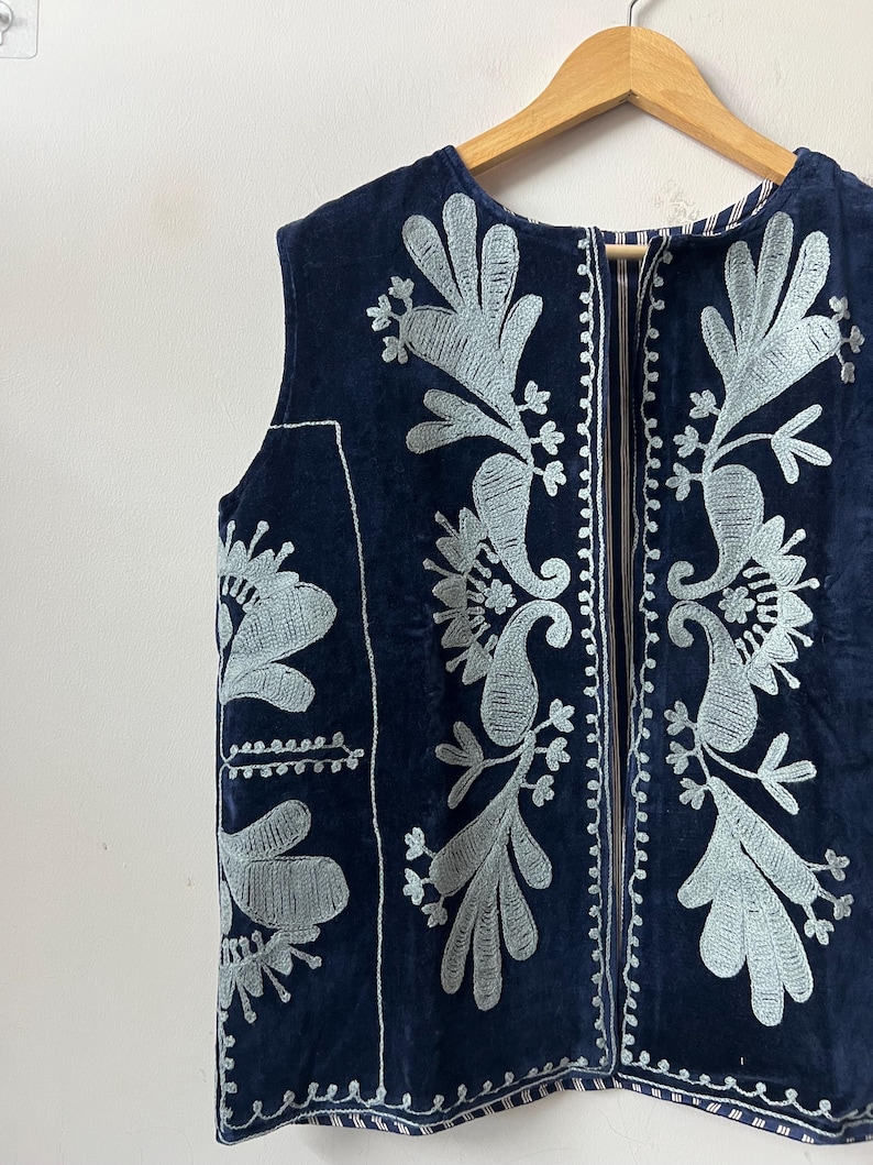 Puede incluir: Un chaleco azul marino con intrincados bordados florales plateados. La prenda sin mangas presenta un cuello redondo y una cremallera vertical. El bordado incluye dise&ntilde;os de cachemir y hojas, creando un aspecto detallado y elegante. El chaleco est&aacute; colgado en una percha de madera.