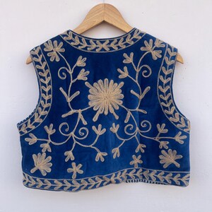 May include: A sleeveless blue vest with intricate gold floral embroidery. The vest is hanging on a wooden hanger against a white background. The embroidery features a central flower design and leafy patterns along the edges.