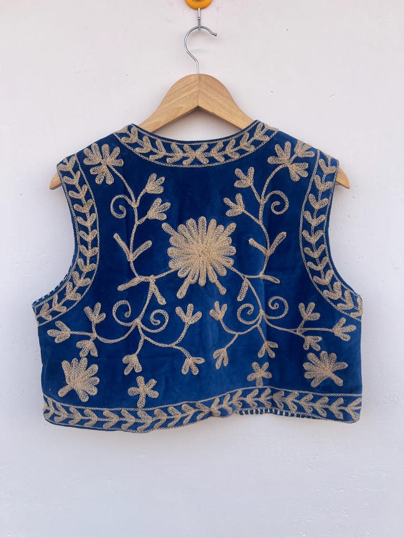 May include: A sleeveless blue vest with intricate gold floral embroidery. The vest is hanging on a wooden hanger against a white background. The embroidery features a central flower design and leafy patterns along the edges.