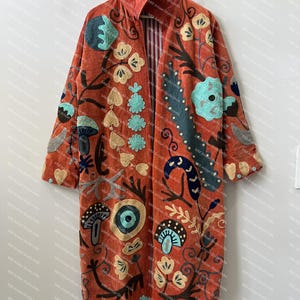 May include: A long, open-front coat in a burnt orange hue, featuring an intricate, multi-colored embroidered pattern of mushrooms, flowers, and abstract botanical shapes in shades of teal, cream, dark blue, and brown, with a striped inner lining.