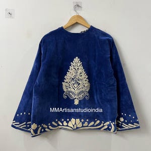 May include: A royal blue velvet jacket featuring cream-colored embroidery. The back displays a large central botanical motif, while the hem and sleeve cuffs are adorned with a repeating floral pattern. The text MMArtisanstudioindia is visible.