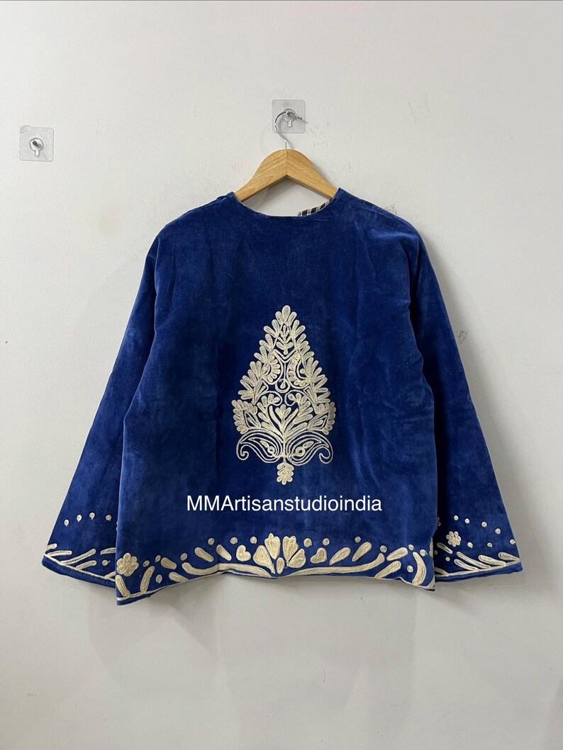 May include: A royal blue velvet jacket featuring cream-colored embroidery. The back displays a large central botanical motif, while the hem and sleeve cuffs are adorned with a repeating floral pattern. The text MMArtisanstudioindia is visible.