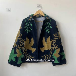 May include: A navy blue jacket with gold and green embroidered bird and leaf designs. The jacket has a striped interior and is hanging on a wooden hanger. The text "MMArtisanstudioindia" is visible on the jacket.
