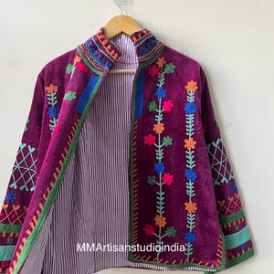 May include: A deep purple embroidered jacket with a high collar, featuring colorful floral and geometric patterns. The jacket has a striped lining and is displayed on a wooden hanger.