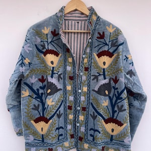 以下が含まれることがあります： 花柄刺繍が施されたライトブルーのベルベットジャケット。 ジャケットはボタン留めで、ストライプの裏地が付いています。 刺繍には、黄色、バーガンディ、緑の花柄のデザインと、青と紫のアクセントが含まれています。 ジャケットは木製のハンガーに掛けられています。