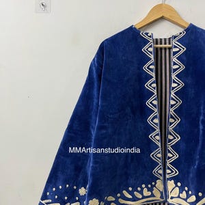 May include: A royal blue velvet jacket featuring cream-colored embroidered trim along the front opening and hem. The embroidery displays geometric and floral patterns. The interior lining is visible, showing a black and white vertical striped fabric.