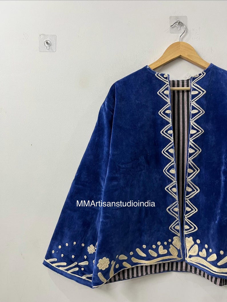 May include: A royal blue velvet jacket featuring cream-colored embroidered trim along the front opening and hem. The embroidery displays geometric and floral patterns. The interior lining is visible, showing a black and white vertical striped fabric.