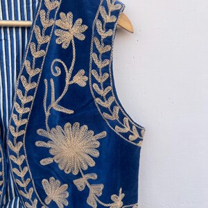 May include: A sleeveless blue vest with intricate beige floral embroidery. The vest features a striped blue and white lining and is displayed on a wooden hanger. The embroidery creates a striking contrast against the deep blue fabric.