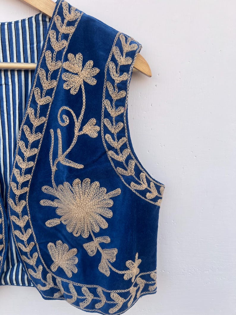 May include: A sleeveless blue vest with intricate beige floral embroidery. The vest features a striped blue and white lining and is displayed on a wooden hanger. The embroidery creates a striking contrast against the deep blue fabric.