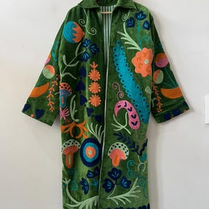 May include: A long-sleeved green velvet coat featuring vibrant, multi-colored embroidery of various floral and mushroom motifs. The coat has a collared neckline, an open front, and a visible white and green striped lining along the inner edges.