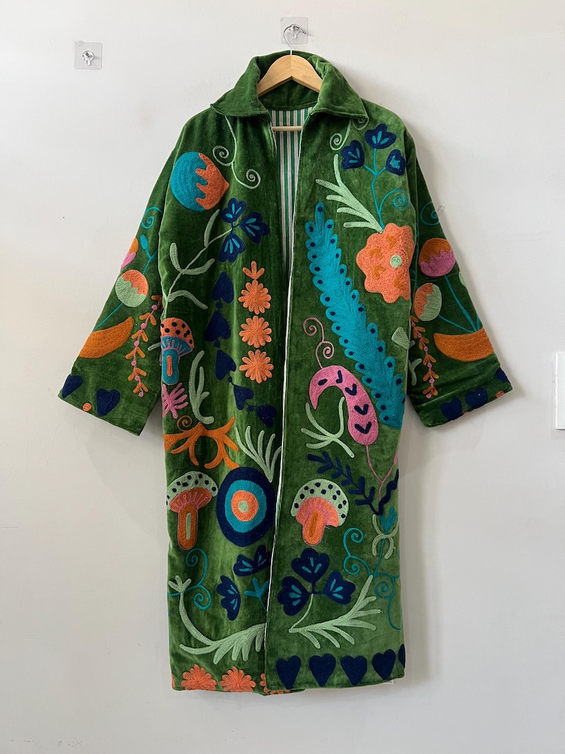 May include: A long-sleeved green velvet coat featuring vibrant, multi-colored embroidery of various floral and mushroom motifs. The coat has a collared neckline, an open front, and a visible white and green striped lining along the inner edges.