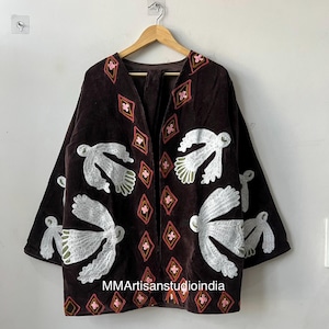 May include: A dark brown velvet jacket with white embroidered bird designs and diamond patterns in pink and red. The jacket has a V-neck and long sleeves. The text "MMArtisanstudioindia" is visible on the bottom of the jacket.