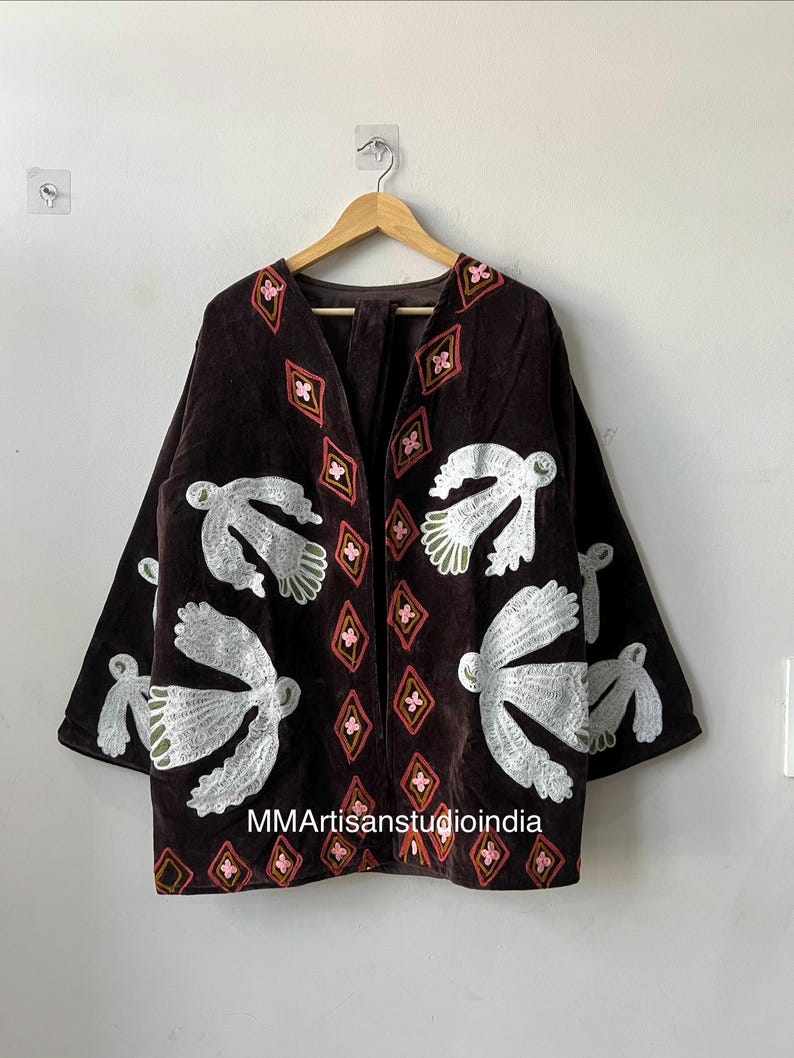 May include: A dark brown velvet jacket with white embroidered bird designs and diamond patterns in pink and red. The jacket has a V-neck and long sleeves. The text "MMArtisanstudioindia" is visible on the bottom of the jacket.