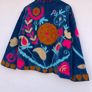 May include: A dark blue velvet jacket with colorful embroidery. The jacket features a high collar and is adorned with floral and heart-shaped designs in shades of pink, gold, and turquoise. The jacket is hanging on a wooden hanger.