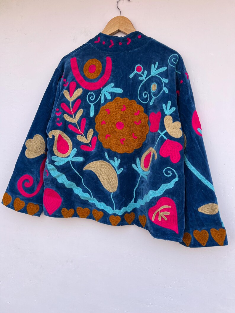 May include: A dark blue velvet jacket with colorful embroidery. The jacket features a high collar and is adorned with floral and heart-shaped designs in shades of pink, gold, and turquoise. The jacket is hanging on a wooden hanger.