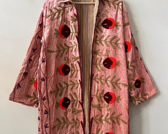 Op de afbeelding: Een lange, open jas in een zachte roze tint, versierd met geborduurde bloempatronen in rood, bruin en groen. De jas heeft een kraag en een gestreepte voering, wat een opvallend contrast creëert. De mouwen zijn lang en de jas hangt aan een houten hanger.