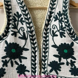 May include: White embroidered vest with dark green floral and leaf patterns. The vest has a cream-colored, fuzzy interior lining. The text "MMArtisanstudioindia" is visible on the bottom of the vest. The vest is hanging on a wooden hanger.