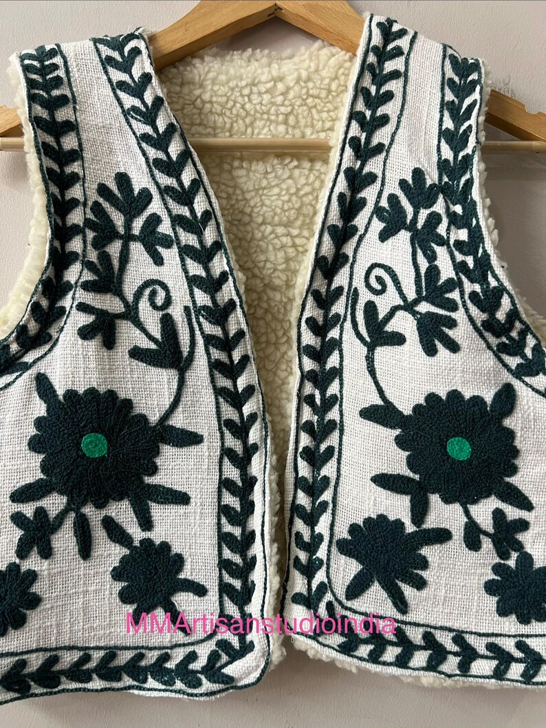May include: White embroidered vest with dark green floral and leaf patterns. The vest has a cream-colored, fuzzy interior lining. The text "MMArtisanstudioindia" is visible on the bottom of the vest. The vest is hanging on a wooden hanger.