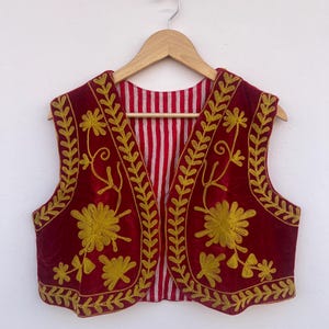 May include: A sleeveless, burgundy vest with gold floral embroidery. The vest has a striped red and white lining and is hanging on a wooden hanger. The embroidery features stylized flowers and leaf patterns.