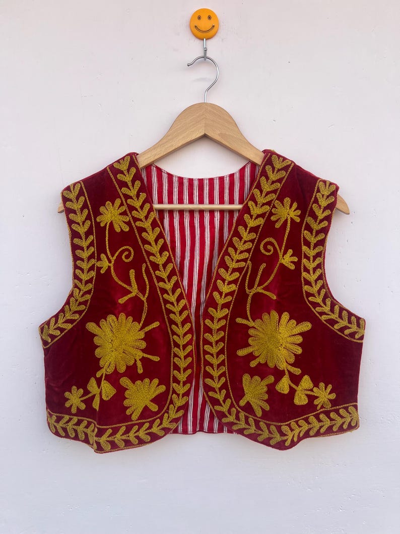 May include: A sleeveless, burgundy vest with gold floral embroidery. The vest has a striped red and white lining and is hanging on a wooden hanger. The embroidery features stylized flowers and leaf patterns.
