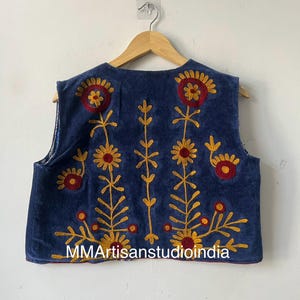 May include: A sleeveless, navy blue vest with embroidered floral designs in red and yellow. The vest features a round neckline and is displayed on a wooden hanger. The text "MMArtisanstudioindia" is visible on the bottom of the vest.