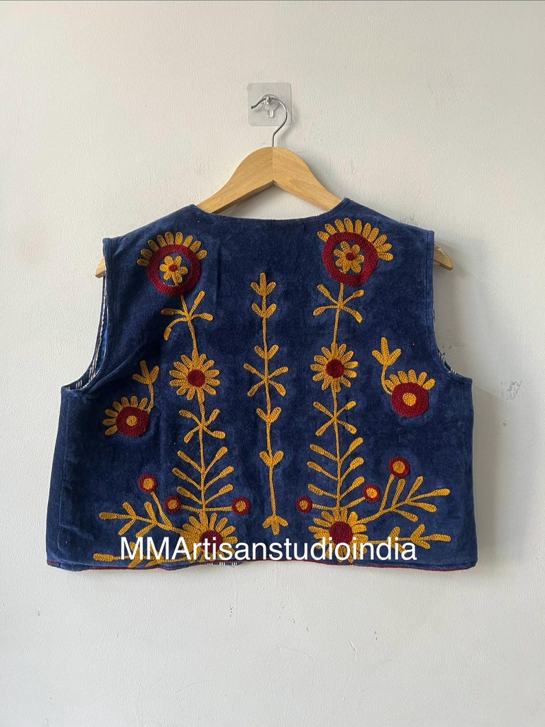 May include: A sleeveless, navy blue vest with embroidered floral designs in red and yellow. The vest features a round neckline and is displayed on a wooden hanger. The text "MMArtisanstudioindia" is visible on the bottom of the vest.