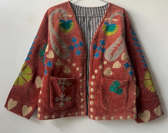 May include: A rust-colored jacket with intricate embroidery. The jacket features a V-neck, two pockets, and a striped lining. The embroidery includes paisley, floral, and heart motifs in shades of green, blue, and beige.