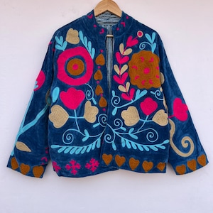 May include: A dark blue velvet jacket with colorful floral embroidery. The jacket features pink, gold, and turquoise embroidered flowers and leaves. The jacket is hanging on a wooden hanger.