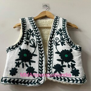 May include: White vest with dark green floral embroidery and a cream-colored sherpa lining. The vest is hanging on a wooden hanger. The embroidery includes floral and leaf patterns. The text "MMArtisanstudioindia" is visible on the bottom of the vest.