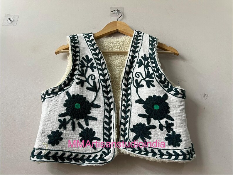 May include: White vest with dark green floral embroidery and a cream-colored sherpa lining. The vest is hanging on a wooden hanger. The embroidery includes floral and leaf patterns. The text "MMArtisanstudioindia" is visible on the bottom of the vest.